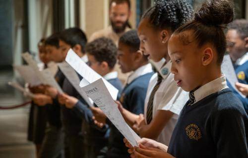 Girls and boys in school uniform sing from sheet music.