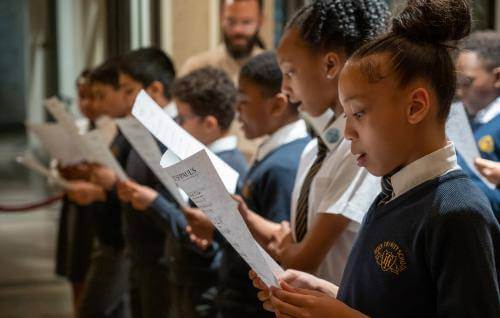 Girls and boys in school uniform sing from sheet music.