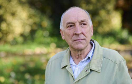 Bob Gilbert is a white man with bald head, wearing a pale green jacket over a blue shirt. He stands outdoors with sun dappled greenery in the blurred background.