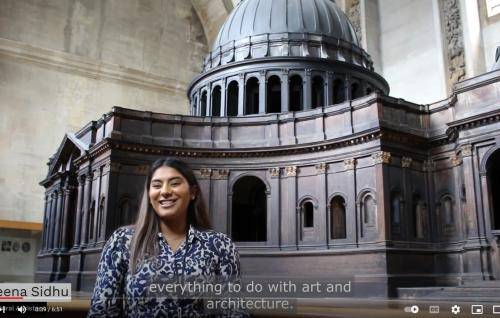 A young Asian woman with long dark hair and wearing a patterned navy and white dress stands in front of a large wooden model of St Paul's Cathedral.