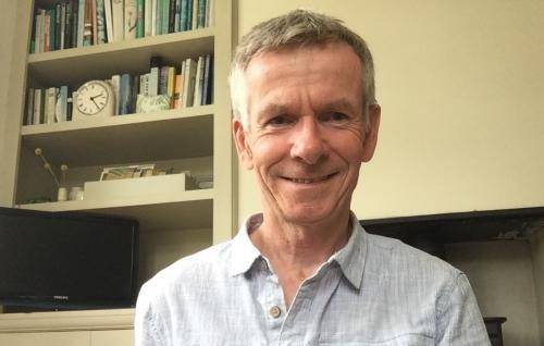 Christopher Chapman is a white man with short grey hair wearing a pair grey casual shirt and sitting in front of a bookcase, cupboard and fireplace.