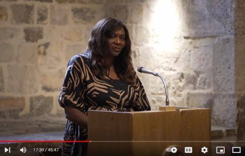 Chine, a black woman with long dark hair wearing a patterned black dress, speaks at the lectern in the Wren Suite of St Paul's Cathedral.