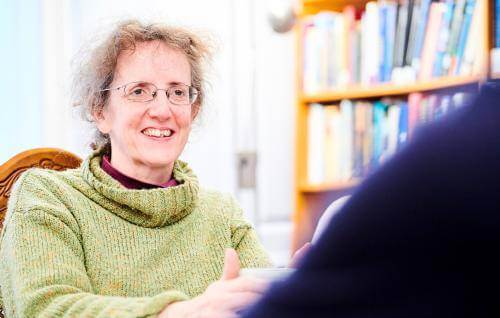 Karen is a white woman with light wavy hair pinned up and glasses, wearing a pale green rollneck sweater, smiling at someone to the side of the camera.