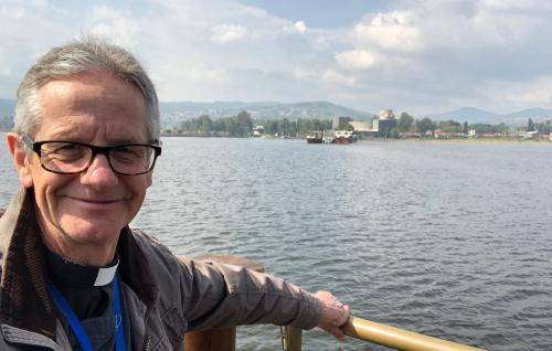 Richard is a white man with short grey hair and black rimmed glasses. He wears a clerical collar and black shirt under a casual light jacket and stands holding onto the wooden railing of a boat or a jetty on a lake. The shoreline in the distance has trees, and some buildings, and there are mountains in the distant background. The sky is pale blue with some clouds and the sun is shining on Richard.