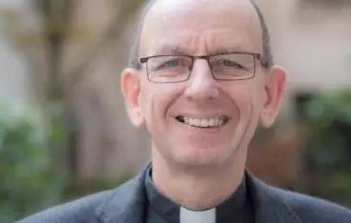 Neil is a white man with balding grey hair and wire framed glasses, wearing a black clerical shirt and black jacket. He smiles at the camera, trees and a garden wall blurred in the background.