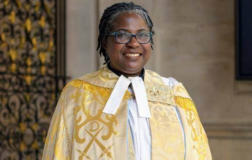 Adéọlá is a black woman with short dark hair, greying around the face. She wears dark blue glasses and a golden embroidered cope with a white preaching band.
