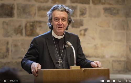 Andrew is a white man with longish grey hair wearing a clerical collar, black suit and a large cross on a chain. He stands at a lectern in front of an old stone wall.
