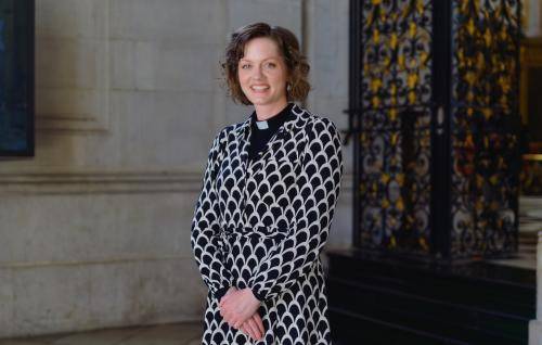 Tessa is a young white woman with wavy brown hair wearing a clerical collar under a black and white patterned dress, standing by the wrought iron gates near the high altar at St Paul's.
