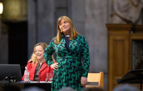 Kate on stage wearing a green dress and clerical collar, with her hand on her hip, giving side eye to the audience as Paula laughs in the background.