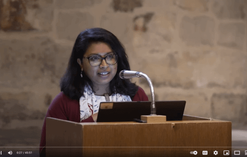 Anupama is a your Asian woman with shoulder length black hair wearing glasses. She is standing at a lectern with a microphone in the Wren Suite at St Paul's Cathedral.