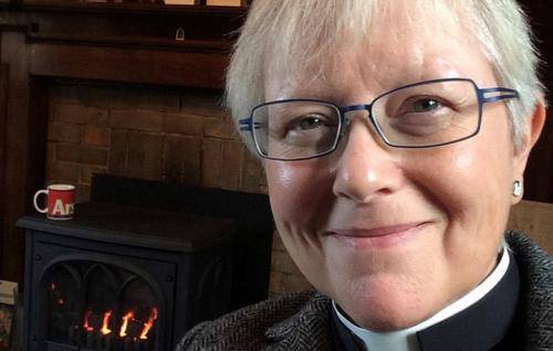 Cally is a white woman with white hair and glasses wearing a clerical collar under a black shirt and grey tweed jacket. She is in front of a mantlepiece decorated with card, framed photos and an Icon, and a lit log burner. She is smiling.