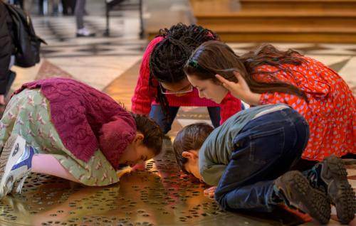 Four young children kneel on the floor of St Paul's Cathedral, with their faces close to the ground, looking through a golden grille through to the Cathedral Crypt.