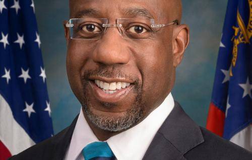 Raphael Warnock is a black man with a bald head, wearing frameless glasses, a dark suit with a white shirt and stripy tie. Behind him are two US flags.