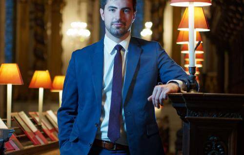 A portrait of James Orford. James is a white man with dark, short hair and a beard. He is wearing a suit and tie, and is stood in the Quire of St Paul's Cathedral.