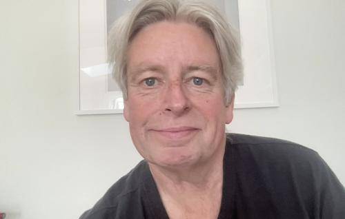 Graeme is a white man with short grey hair and a black top, seated in front of a white wall with a white framed photo behind him.