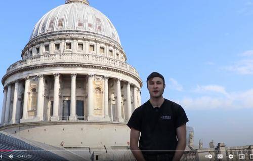 A young white man wearing a black cap and a black St Paul's t-shirt stands on the roof of the cathedral, the dome in the sunshine behind him.