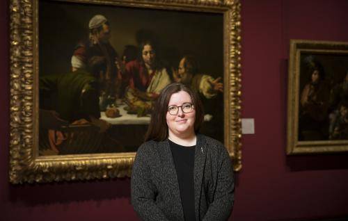 Siobhan Jolley is a white woman with long dark hair and round glasses, wearing a dark grey jacket over a black dress. She stands in the National Gallery in front of Caravaggio’s painting The Supper at Emmaus.