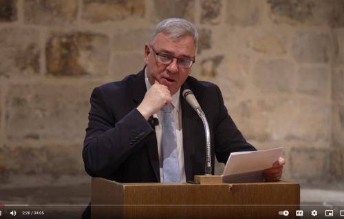 Andrew Carwood, a white man with grey hair and glasses, in a dark suite with a pale blue tie, stands at a microphone in front of a stone interior wall