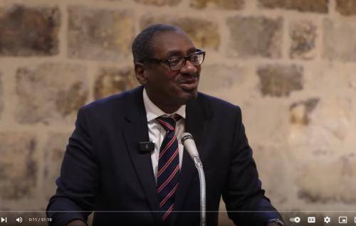 Richard is a black man with glasses wearing a navy suit and tie talking at a microphone in front of a stone wall