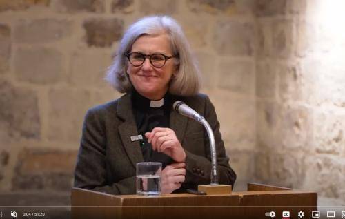 Janet is a white woman with grey bobbed hair and black rimmed glasses wearing a clerical collar and black shirt under a dark grey jacket, standing in front of a lectern and microphone and an interior stone wall