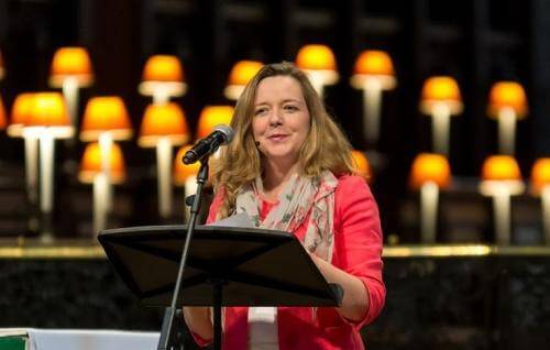 Paula is a white woman with long blond hair wearing a red jacket and grey scarf and standing in front of a microphone and music stand with the glowing lamps of the quire of St Paul's Cathedral in the background