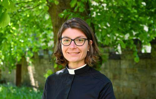 Stephanie is a white woman with shoulder length dark hair wearing dark rimmed glasses and a clerical collar under a black cassock, standing in front of a row of leafy green trees.