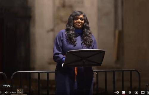 Chine is a black woman with long curly black hair wearing a dark blue dress and standing at a lectern in St Paul's Cathedral