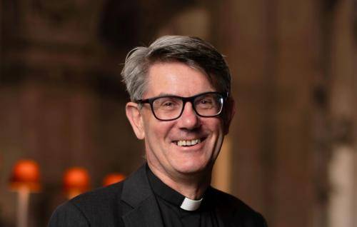 Andrew is a white man with short grey hair and black rimmed glasses wearing a clerical collar, black shirt and black jacket. The lamps of the quire of St Paul's in the background.