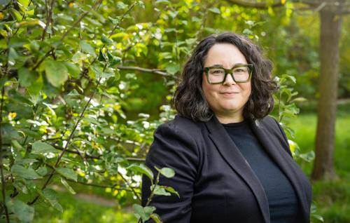 A white woman in clerical collar wearing dark green glasses standing in front of sun dappled trees.