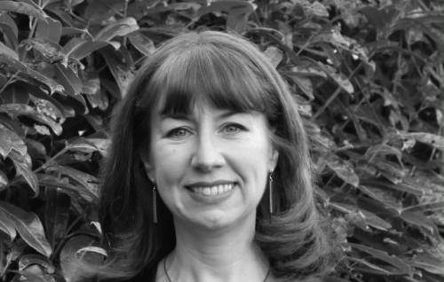 Anna is a white woman with shoulder length dark hair standing in front of a leafy hedge