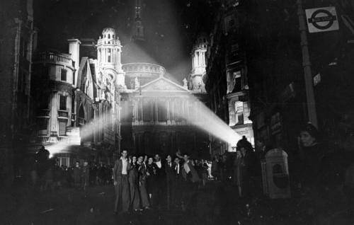The West End of St Paul's, lit by searchlights, with a crowd of people posing for a photo in front of it.