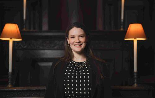 A photo of Hilary Punnet, Assistant Director of Music at St Paul's Cathedral. Hilary is a white woman with long brown hair. She is smiling at the camera, and wearing a black polka dot dress with a black blazer.