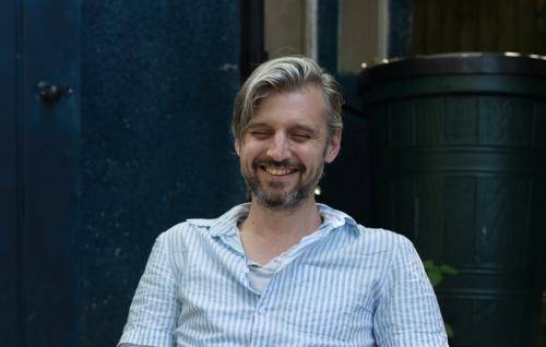 David is a white man with light brown hair and a darker beard, sitting outside in a garden chair, wearing a pale blue short sleeved shirt.