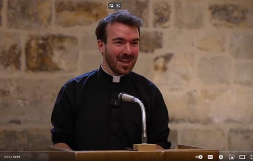 Will is a white man with short dark hair and beard, wearing a black clerical shirt and white collar, standing at a microphone in the Wren Suite at St Paul's Cathedral