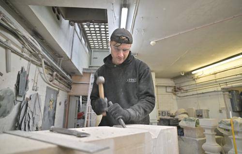 male person wearing a cap and black clothes in a workshop using a hammer and chisel to carve a stone