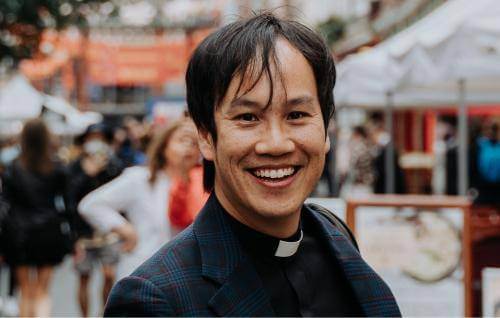 Mark Nam headshot wearing a clerical collar under a black suit and shirt, standing in a busy marketplace