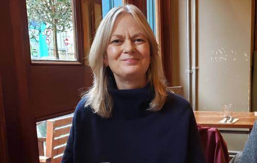Linda wears a navy polo neck jumper and sits in a cafe by windows overlooking a tree in a city