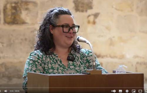 Karen wears glasses and a green and white patterned dress and stands at a lectern with microphone