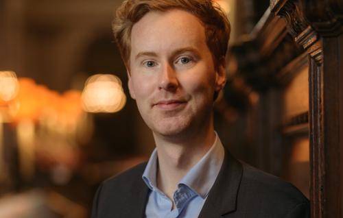 James stands in the quire of St Paul's Cathedral wearing a black jacket over a pale blue shirt