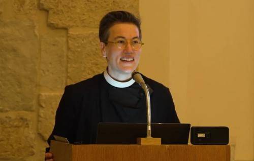 Ayla wears a clerical collar and black shirt and stands at a microphone and lectern
