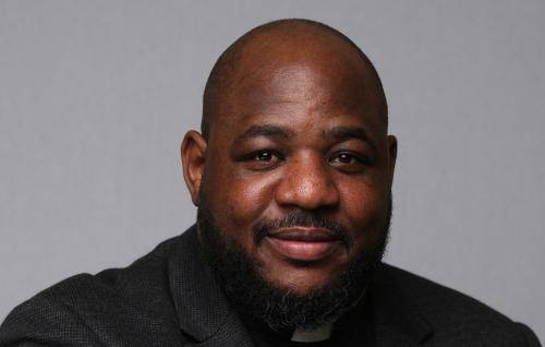 Carlton wears a clerical collar with a black shirt and jacket