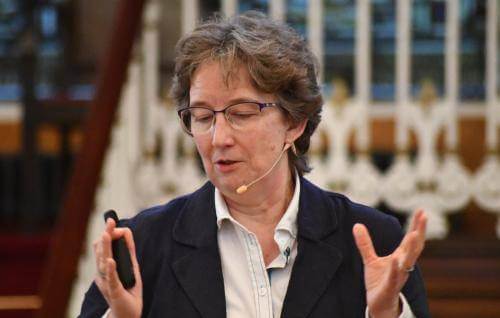 Helen has short brown hair and glasses, and stands with hands raised and wearing an ear microphone as she talks.