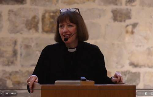 Carys Walsh, wearing a black shirt and clerical collar, with an ear microphone and glasses on her head, stands at a lectern in the Wren Suite at St Paul's Cathedral.