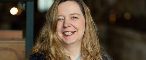 Paula Gooder is a white woman with long blond hair wearing a dark jacket and blue pendant necklace. She smiles to the camera and is standing indoors with the blurry glow of windows behind her in what appears to be a very large space. 