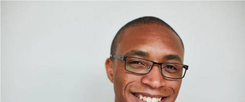 Jarel is a black man with shaved head add black framed glasses, wearing a cerical collar with a black shirt and jacket. He smiles to the camera before a pale background.