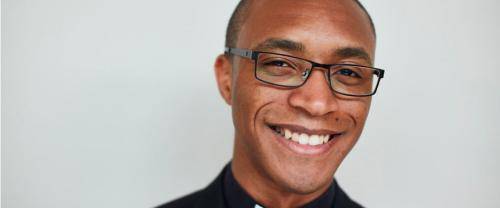Jarel is a black man with shaved head add black framed glasses, wearing a cerical collar with a black shirt and jacket. He smiles to the camera before a pale background.