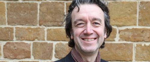 Andrew is a white man with dark hair streaked with grey, wearing a black jacket and burgundy scarf, standing in front of an old brick wall, smiling to the camera.