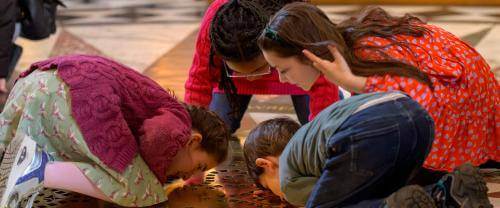 Four young children kneel on the floor of St Paul's Cathedral, with their faces close to the ground, looking through a golden grille through to the Cathedral Crypt.