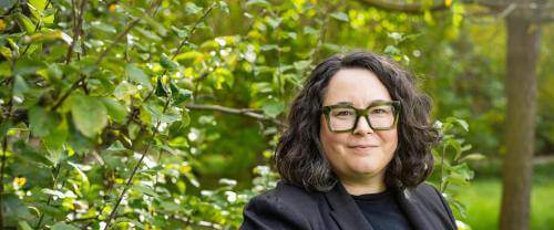 A white woman in clerical collar wearing dark green glasses standing in front of sun dappled trees.