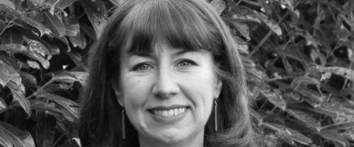 Anna is a white woman with shoulder length dark hair standing in front of a leafy hedge